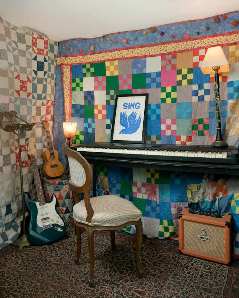 Cozy music corner with a piano, striped chair, colorful quilted wall, guitar, and lamp.