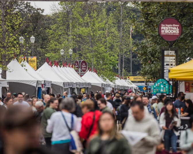 About a hundred people, mostly out of focus, walk to and from white book seller tents on a tree-lined college campus. In the background, a sign reads: "Los Angeles Times Festival of Books."