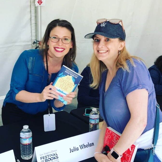 Two women with light skin tone and long hair--one a brunette with reading glasses, the other a blonde with her reading glasses resting on her head--smile at a book signing table with a placard that reads "Julia Whelan." The brunette is holding a book titled "My Oxford Year."