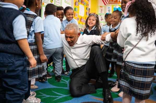 Former President Barack Obama pretends to struggle to stand as children lift him up during a visit to Learning Through Play Pre-K with Mayor Zohran Mamdani in the Bronx in New York, Saturday, April 18, 2026. (AP Photo/Angelina Katsanis)