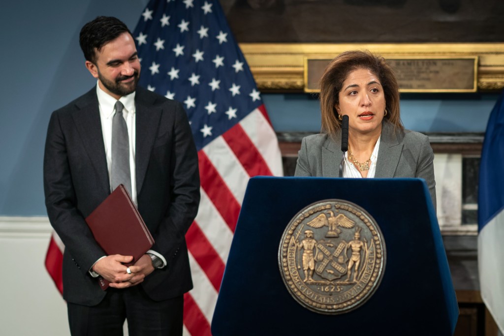 Nadia Shihata speaks at a podium wearing a grey suit during her nomination as DOI commissioner. Mayor Mamdani stands on the left, smiling, wearing a black suit and grey tie.