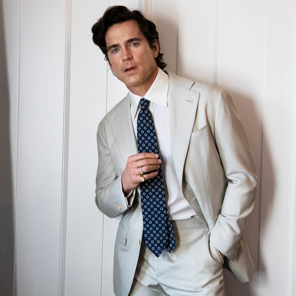 A man dressed in a light gray suit and tie stands against a wall.