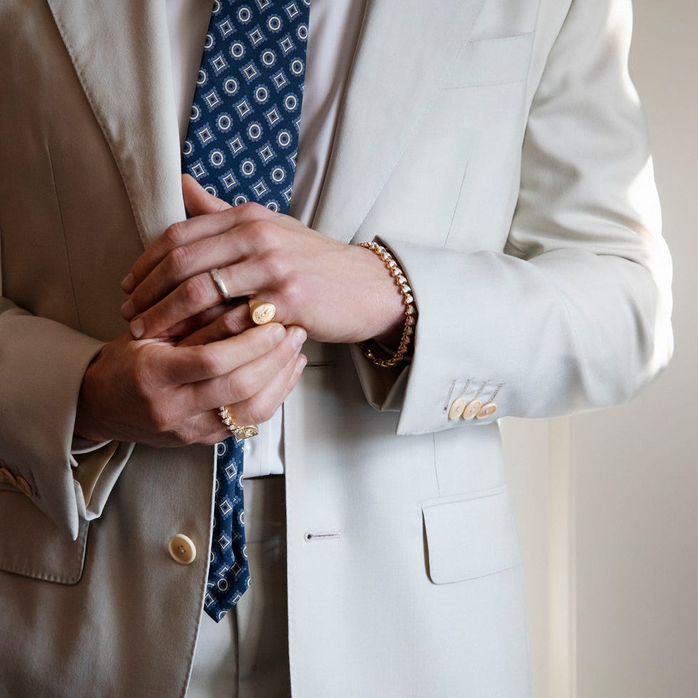 A man wearing a light suit and patterned tie, adjusting jewelry on his hands.