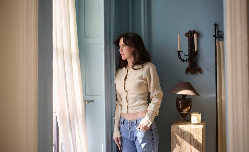 A woman in a cream cardigan and blue jeans stands by a window, with a lamp and candle on a side table.