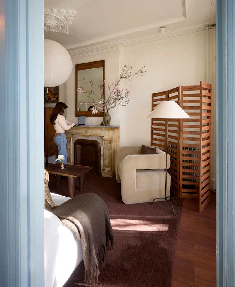 Cozy living space with a woman using a laptop by a vintage fireplace, a wooden screen, and a floral arrangement.