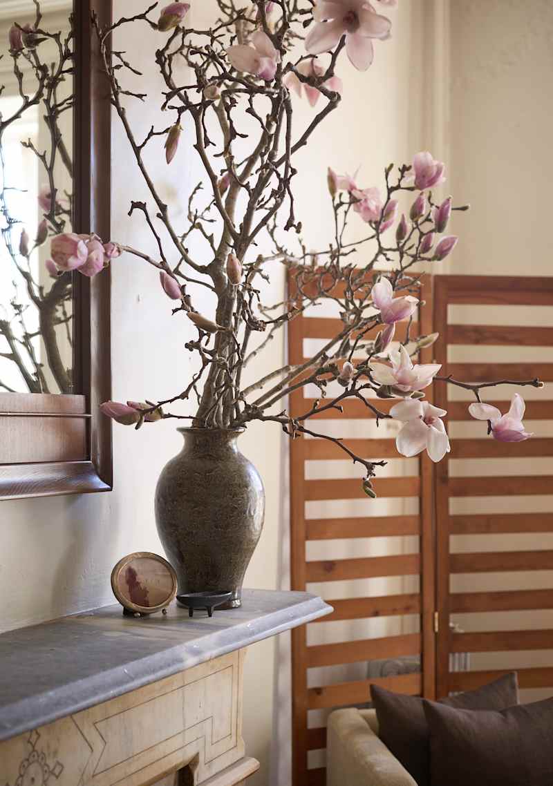 Magnolia branches with pink flowers in a dark vase on a mantel, with a round decorative frame and wooden screen in the background.