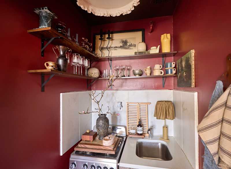 Cozy kitchen corner with red walls, wooden shelves displaying glassware and pottery, and a small sink with decor.