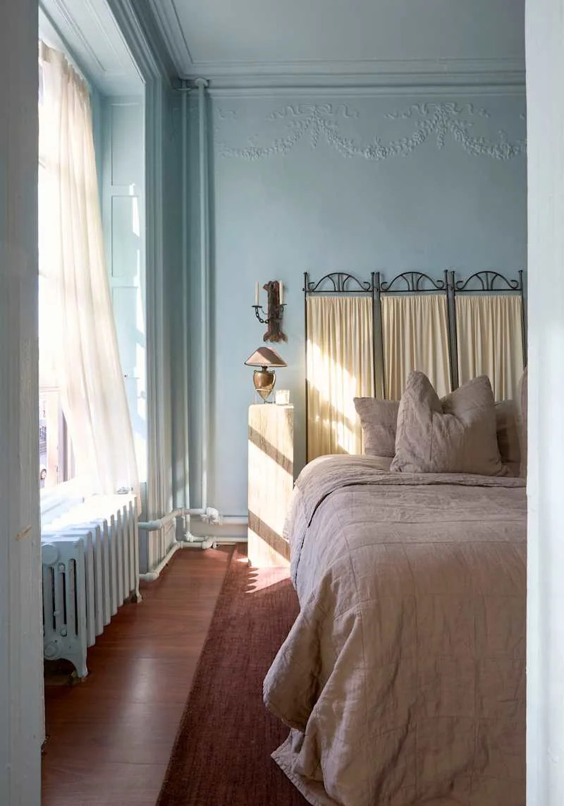 Cozy bedroom with a light blue wall, a bed with beige bedding, a wooden nightstand, and a vintage lamp.