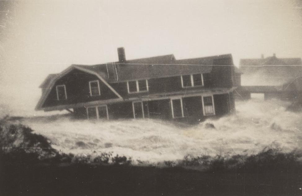 A black-and-white photo shows a two-story house carried away by waves