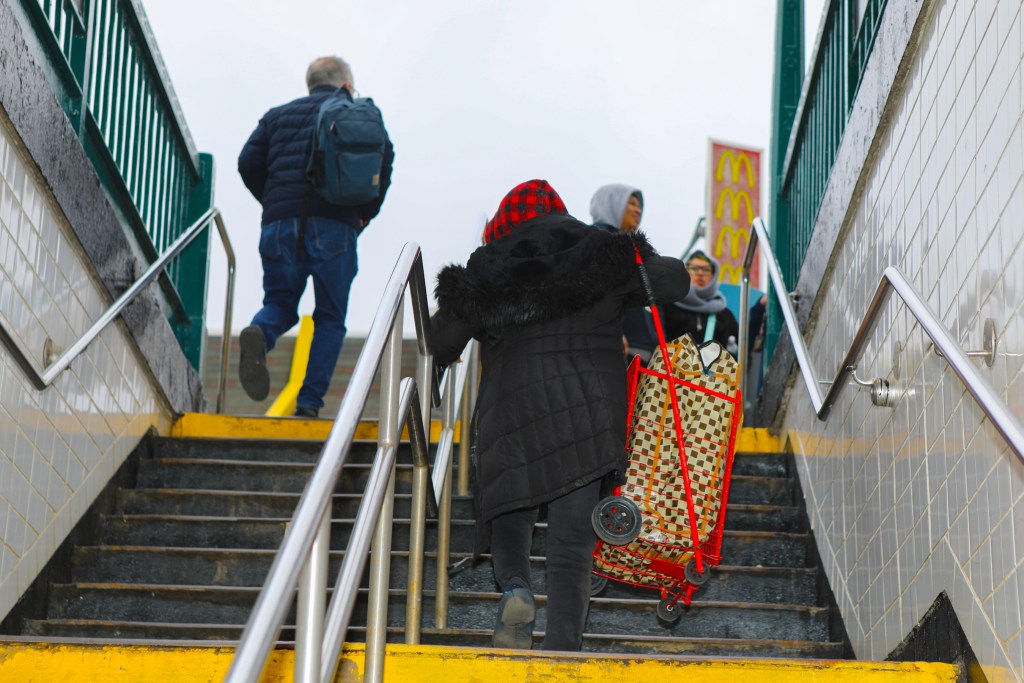A woman holds the subway station stairway rail in one hand and a full red grocery cart in the other.