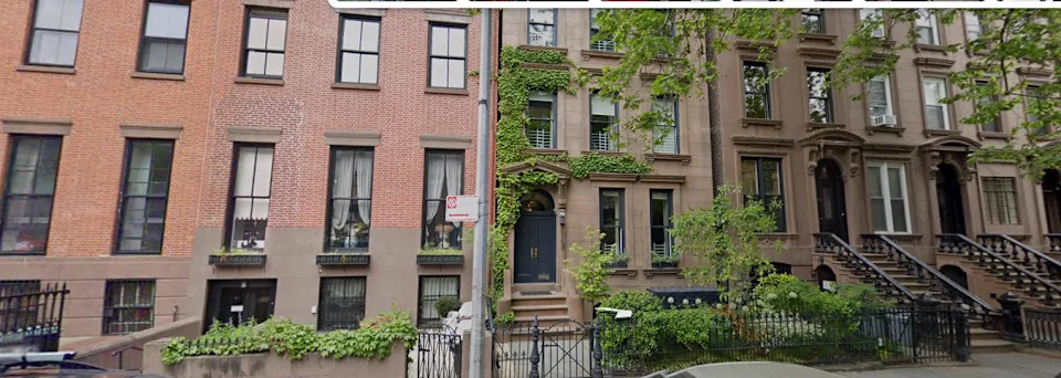 Row of brownstone buildings with greenery and a sidewalk.