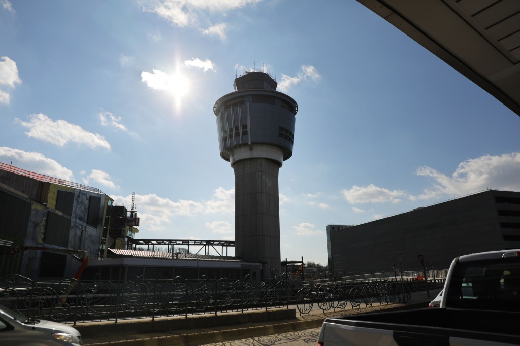 A control tower at LaGuardia Airport, with buildings and parked cars, under a blue sky with some clouds.