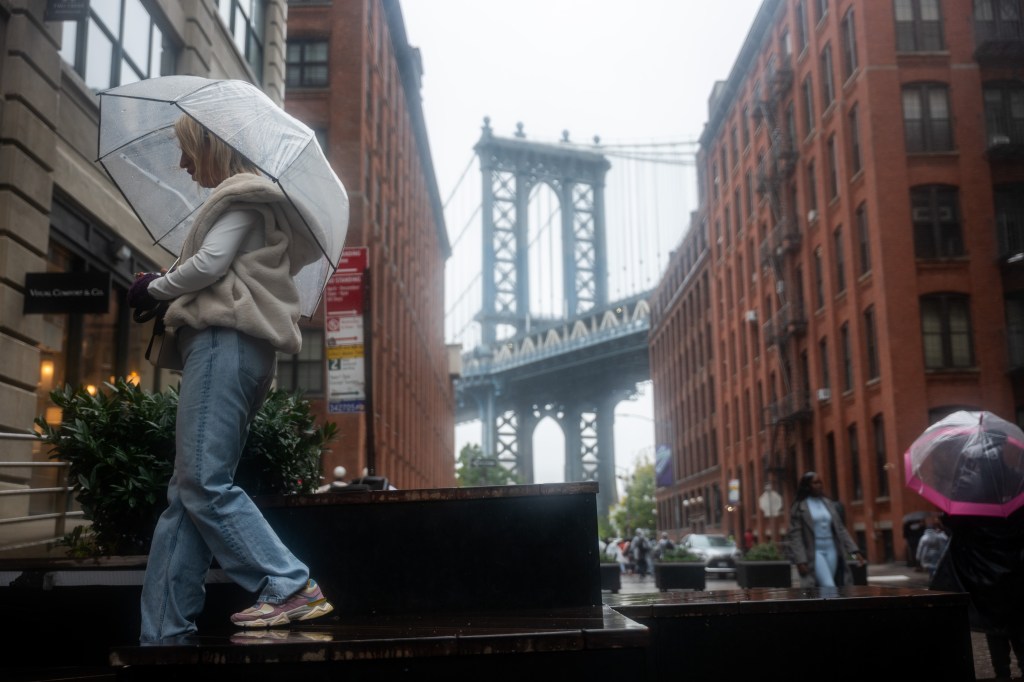 A woman with a clear umbrella walks in the rain in Brooklyn with the Manhattan Bridge in the background.