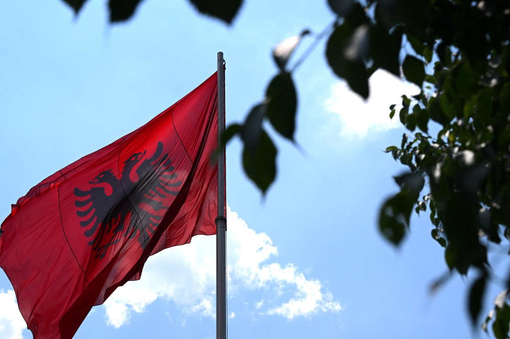 The Albanian flag, red with a black double-headed eagle, flying against a blue sky with clouds.