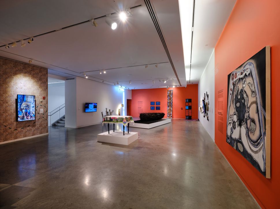 A wide view of a contemporary art gallery at the Bronx Museum with paintings, sculptures and video works displayed across white and orange walls for an article about its current programming during renovation.