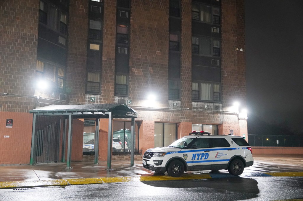 A white NYPD police vehicle with its red and blue lights illuminated is parked in front of a brick apartment building at night.
