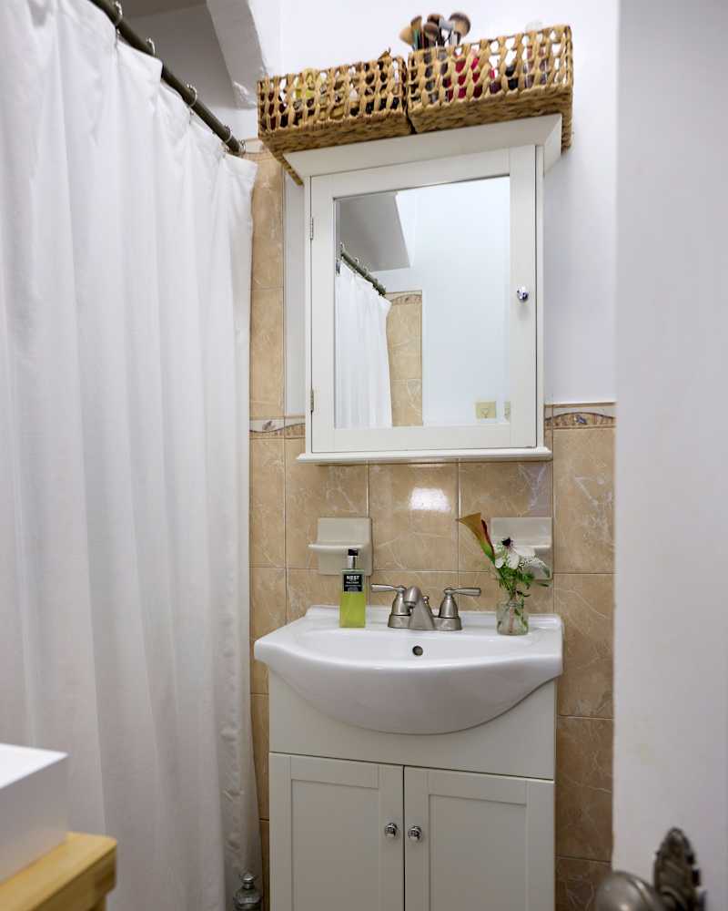 Small bathroom with a white sink, silver faucets, a mirror, and a basket of makeup brushes above.