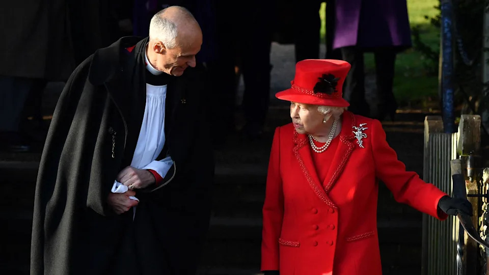The late Queen is on the right. She is wearing a red dress, a red coat and a red hat. She is holding a railing on the right and speaking to a reverend, who is on the left. He is leaning towards her.