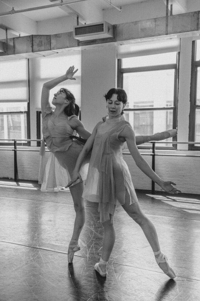 In this black and white photo, two women in filmy costumes pose next to each other. One does a piqué arabesque on pointe, resting her left hand on the other woman's right shoulder. The standing woman poses in tendu dérriére, her amrs in mid-motion.