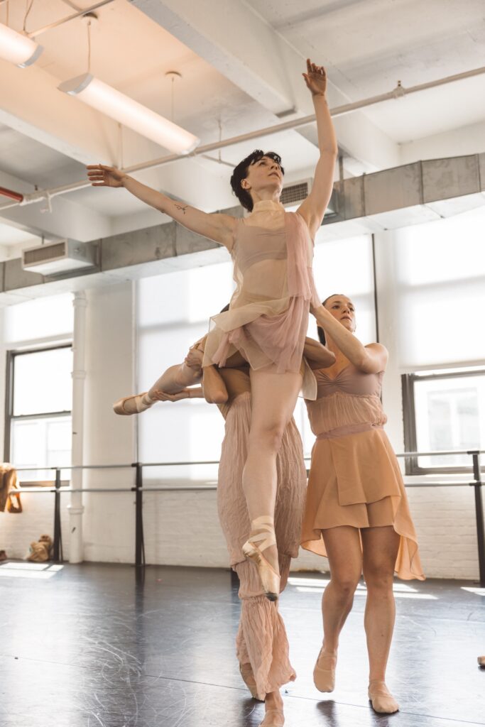 Three women dancers in filmy neutral-colored costumes do a partnered lift. One woman in arabesque is lifted onto the shoulder of another, while the third walks closely beside them and supports the lifted dancer's waist.