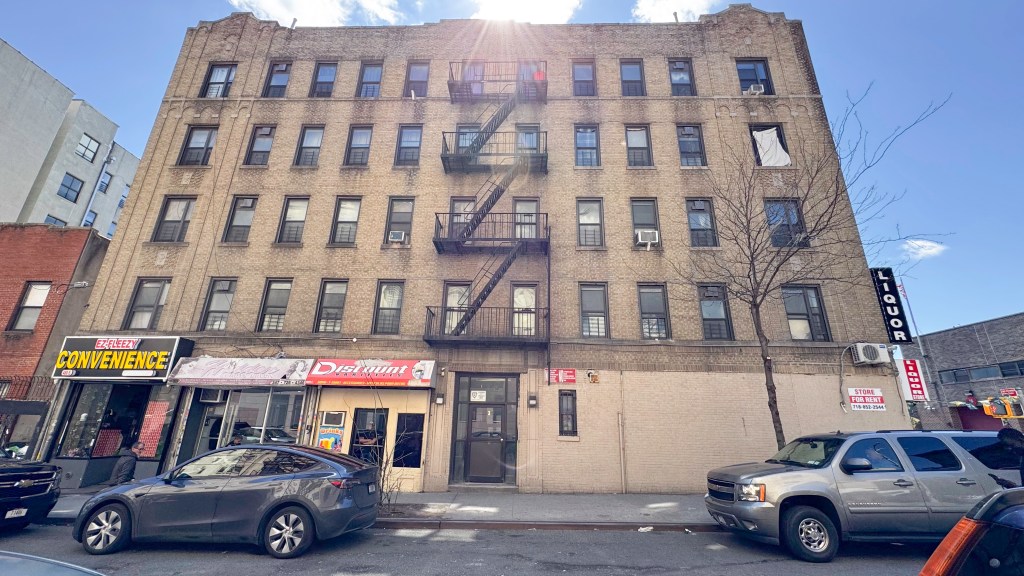 A tall, light brown brick apartment building in the Bronx with a fire escape and a "LIQUOR" sign on the right.