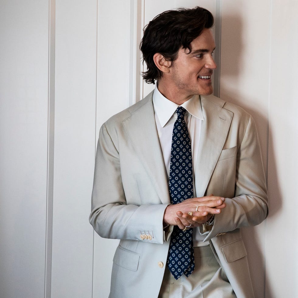 A person in a light-colored suit and tie, standing against a wall.