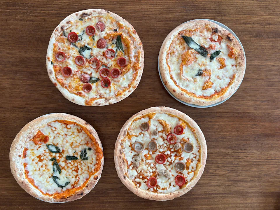 Four pizzas on a wooden table, each topped with various ingredients including pepperoni, sausage, and basil
