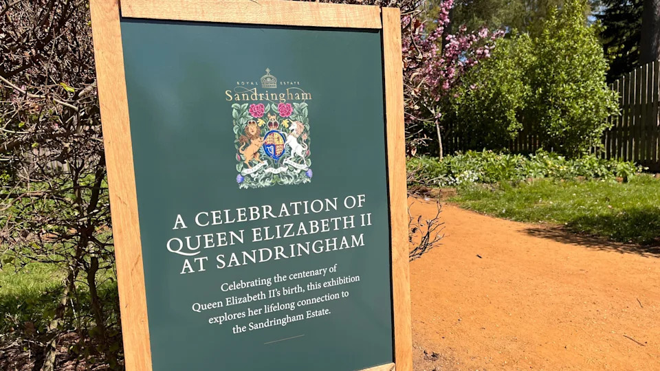 An information board is on the left of the picture. It is beside a path. There are trees and shrubs in the background. The board is green and has a wooden frame. It reads: "A celebration of Queen Elizabeth II at Sandringham."