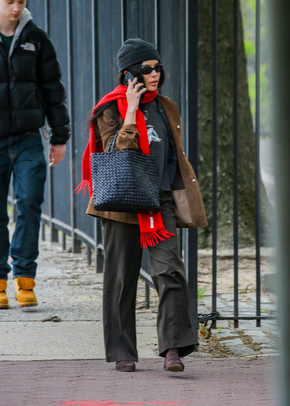Person walking on a city sidewalk, talking on the phone, wearing a red scarf.