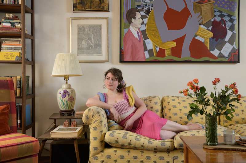 A young woman in a pink outfit lounges on a yellow patterned sofa, surrounded by bookshelves and artwork.