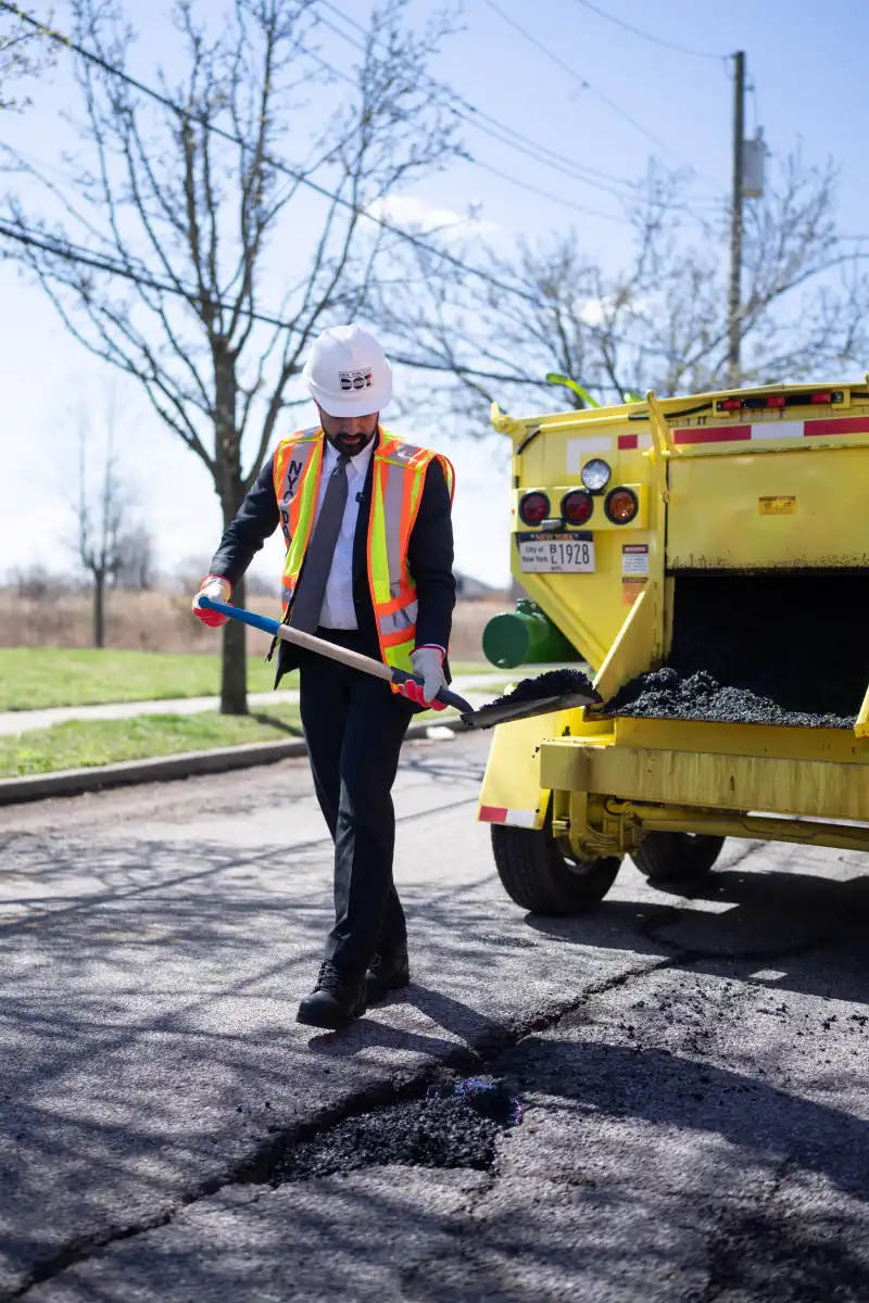 mayor mamdani holding shovel full of asphalt while fixing pothole on street