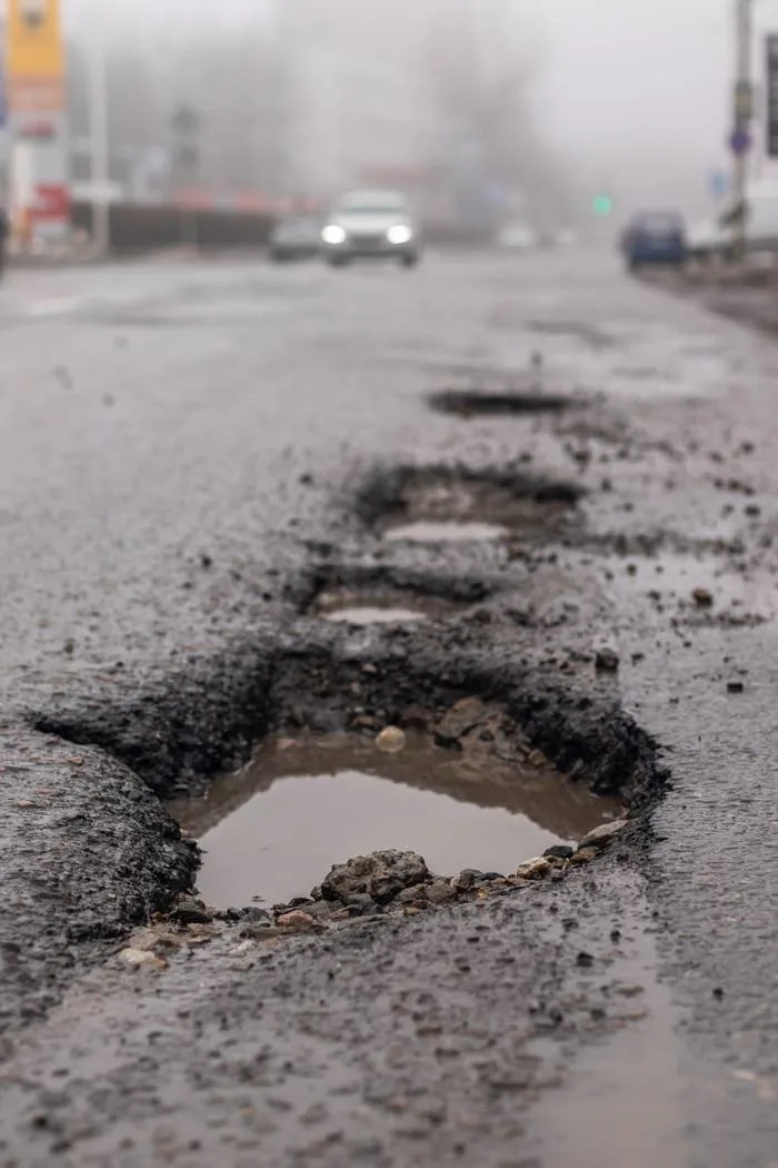 Pothole-filled road with visible water accumulation, leading into the distance on a foggy day, with blurred car in the background