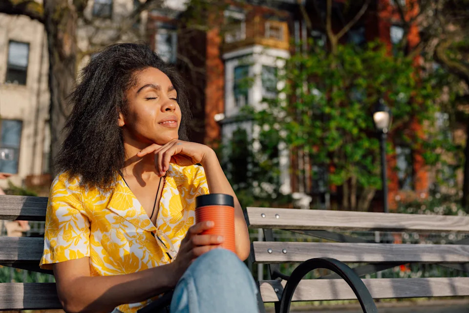 A woman relaxes on a bench, tilting her head back to catch the sunlight. She is holding a coffee cup and wearing a bright yellow summer shirt.
