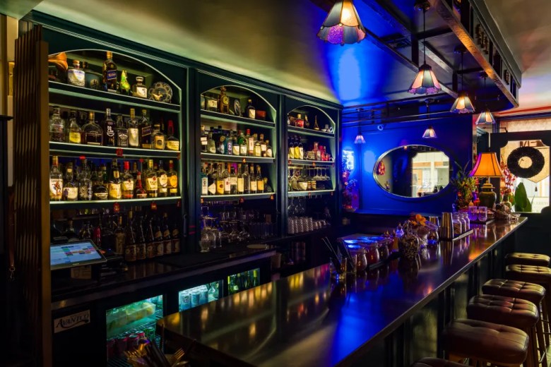 Dimly lit cocktail bar interior with floor-to-ceiling liquor shelves, Tiffany pendant lamps, blue neon accent lighting, and leather bar stools.
