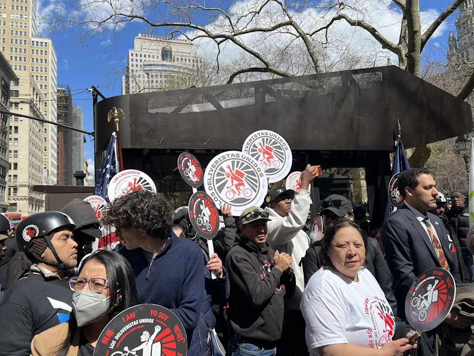 A group of app-based delivery workers hold up signs for their union, Los Deliveristas Unidos, while standing next to New York City's first-ever rest stop for delivery workers. The hub is an industrial-looking, metallic structure. There is a tree and a blue sky with some clouds in the background, and some buildings to the left.
