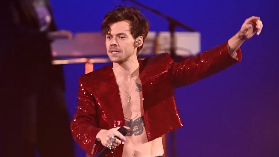 Harry Styles performing on stage, wearing a red sequin jacket, holding a microphone, with tattoos visible on his chest.