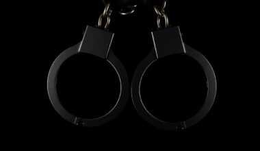 An extreme close-up photograph of a pair of metal handcuffs against a completely black background, the harsh lighting creating dramatic shadows and textures to convey the gravity of the alleged crimes.