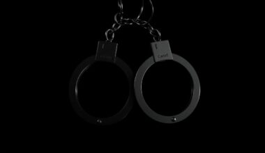 An extreme close-up photograph of a pair of handcuffs against a pitch-black background, creating a stark, gritty, investigative aesthetic through the use of harsh, direct camera flash lighting.