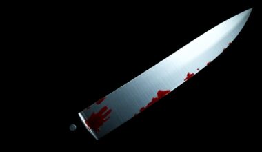 An extreme close-up of a single bloody knife blade, captured in stark contrast against a pitch-black background using harsh, direct flash photography to create a gritty, investigative aesthetic.