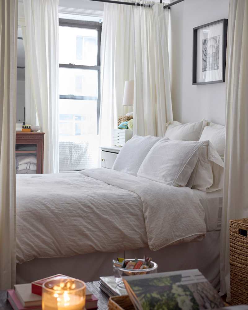 Cozy bedroom with a white bed, sheer curtains, a lamp, and a wooden dresser, featuring books and a candle on a nightstand.