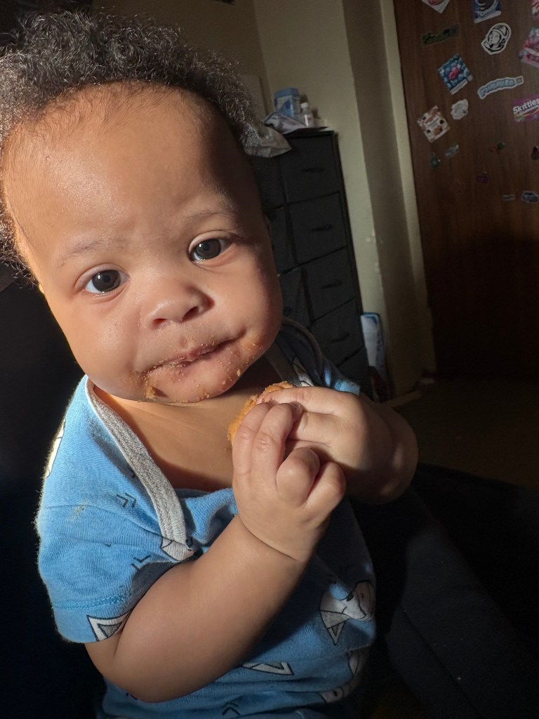 A 7-month-old girl with dark curly hair and a light blue onesie sits facing the camera, her mouth smeared with food, holding a piece of food in her hands.