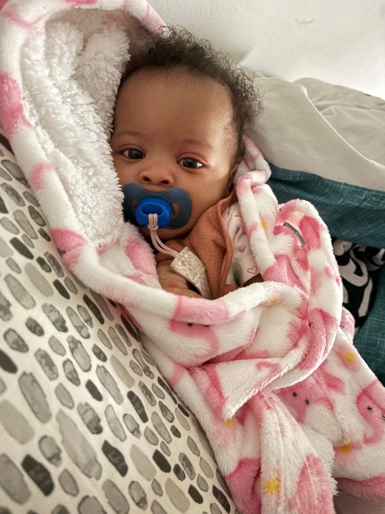 A baby with dark curly hair and dark eyes looking at the camera, wearing a blue pacifier, wrapped in a pink and white blanket.