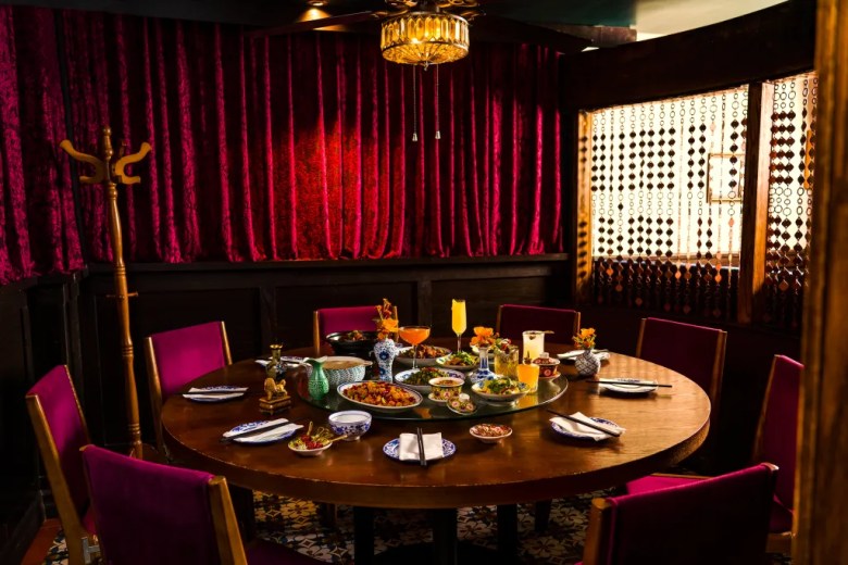 Elegant private dining room with lazy Susan table set with Chinese dishes, cocktails, velvet curtains, and jewel-toned chairs.