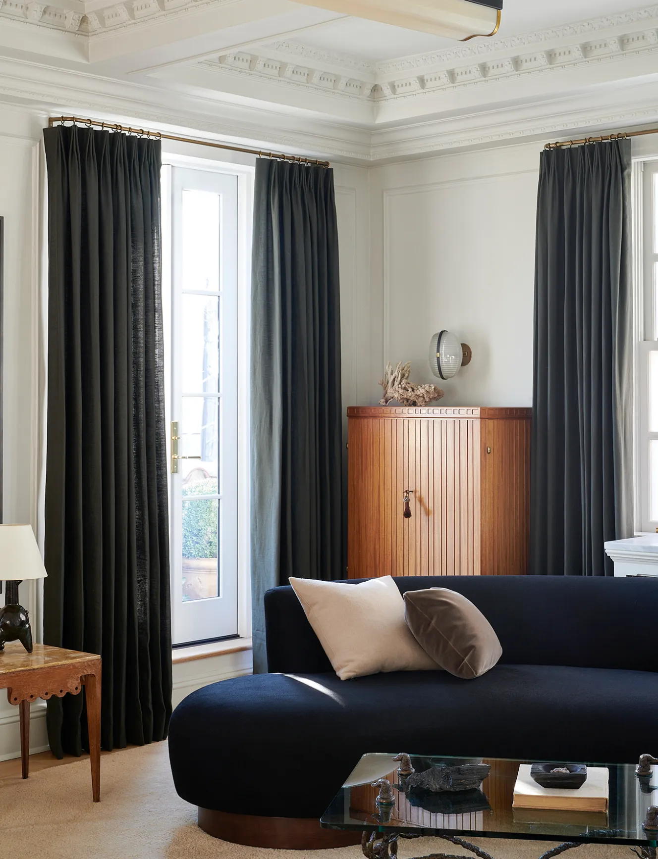 A living room with a black curved sofa, a glass coffee table, and dark gray curtains framing a white french door.