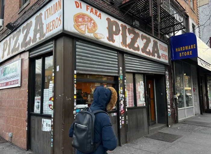 Person walking past Di Fara Pizza storefront with its signage visible; adjacent is a hardware store