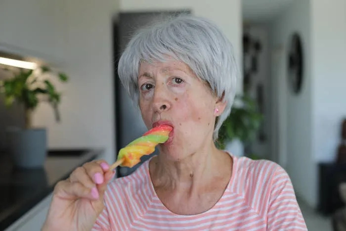 Older woman with short grey hair enjoying a colorful popsicle in a casual, striped top inside a home setting