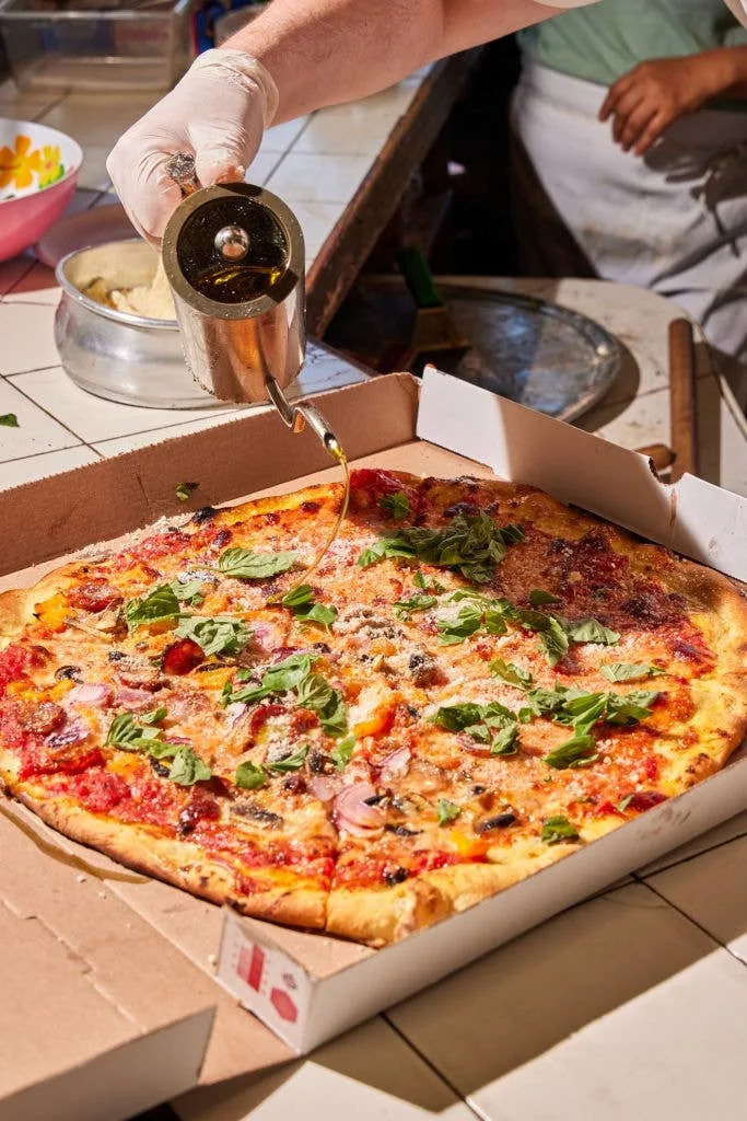 Person pouring oil over a pizza in a takeout box, topped with basil, mushrooms, and other ingredients