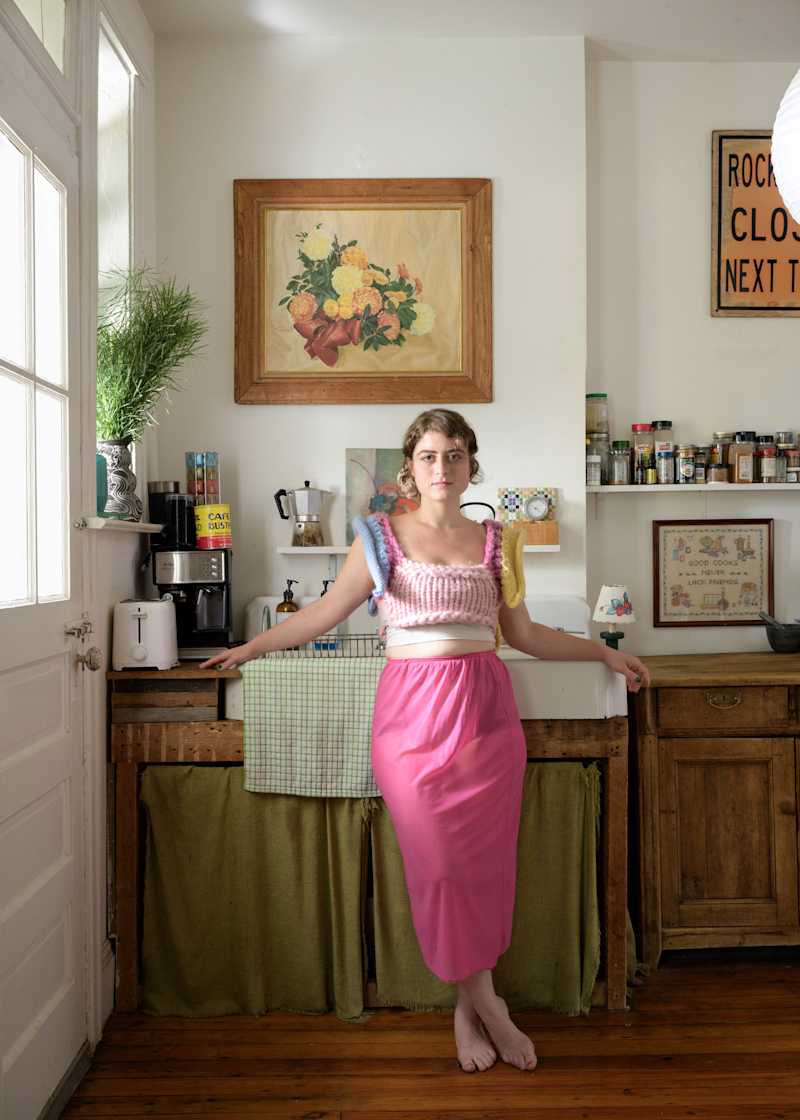 A woman in a pink skirt and colorful knitted top stands in a bright kitchen with wooden accents, plants, and artwork.