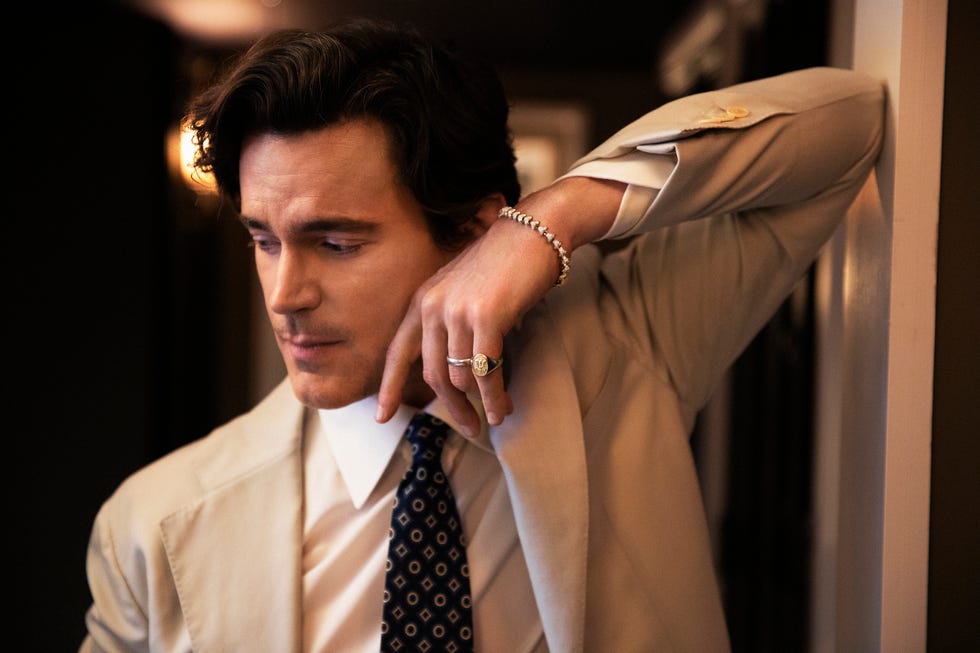 Person leaning against a wall, wearing a suit and accessories.