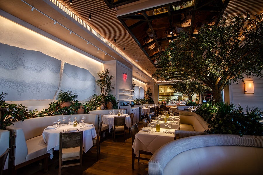 A stylish restaurant interior featuring white tablecloth-covered tables set with wine glasses, plates, and cutlery. The seating includes cushioned booths and wooden chairs. The space is decorated with lush green plants and trees, creating a natural ambiance. The ceiling has wooden slats with recessed lighting, and the walls have modern art pieces and soft lighting. The overall atmosphere is warm and inviting.